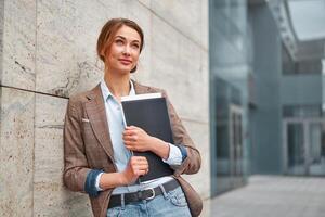 blond Geschäft Frau mit ein Mappe von Unterlagen im ihr Hände Träume und sieht aus hoch. verschwommen Bild von ein Geschäft Center im das Hintergrund. foto