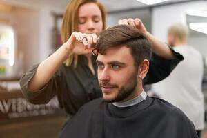 gut aussehend Blau Augen Mann Sitzung im Barbier Geschäft. Friseur Friseur Frau Schneiden seine Haar. weiblich Barbier. foto