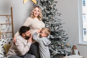 Weihnachten Familie Glück Porträt von Papa, schwanger Mama und wenig Sohn Sitzung Sessel beim Zuhause in der Nähe von Weihnachten Baum Umarmung Lächeln foto