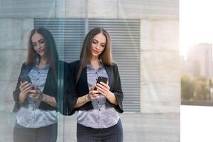 Geschäft Frau 35 Jahre gekleidet Streifen Hemd und schwarz Jacke mit lange Haar Stehen in der Nähe von Büro Gebäude draussen verwenden Smartphone foto