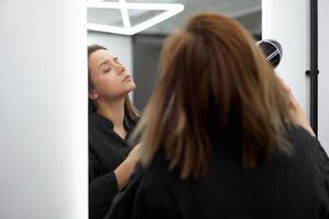 Porträt von ein schön kaukasisch Frau Wer steht in der Nähe von das Spiegel und trocknet Haar mit ein Fön. foto