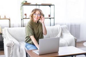 Frau Brille mit Laptop Computer während Sitzung Sofa mit groß Fenster auf Hintergrund beim Zuhause Innere foto