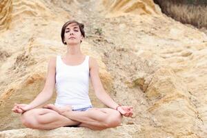 ein schön jung Mädchen mit kurz Haar ist gekleidet im kurze Hose und ein Weiß Jersey ist üben Yoga auf das Hintergrund von Felsen. Pose von das Lotus. das Konzept von Ruhe und Konzentration foto