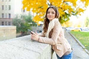 schön kaukasisch Brünette Mädchen Stehen warm Herbst Tag mit Hintergrund von Bäume mit Gelb Laub und ein Stadt. gekleidet wolle Sweatshirt und Jacke. foto