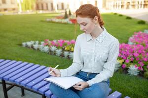ein schön jung rothaarig Mädchen schreibt ein romantisch Liebe Brief Sitzung auf ein Bank in der Nähe von das Institut. Schüler Liebe. oder Hausaufgaben foto