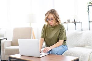 Frau Brille mit Laptop Computer während Sitzung Sofa mit groß Fenster auf Hintergrund beim Zuhause Innere foto