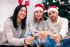 glücklich freunde feiern Neu Jahr im Zuhause Innere im Weihnachten Hüte Sitzung in der Nähe von ein Weihnachten Baum mit Brille von Wein foto