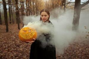 ein schön Mädchen mit lange Haar im ein schwarz Mantel ist halten ein Orange Kürbis von welche Rauch ist Kommen. glücklich Halloween. dunkel Herbst Wald. Schrecken foto