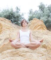 ein schön jung Mädchen mit kurz Haar ist gekleidet im kurze Hose und ein Weiß Jersey ist üben Yoga auf das Hintergrund von Felsen. Pose von das Lotus. das Konzept von Ruhe und Konzentration foto