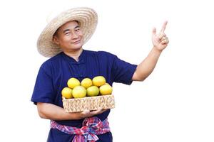 gut aussehend asiatisch Mann Farmer trägt Hut, Blau Shirt, hält Korb von organisch Orange Früchte, Punkt Finger hoch, isoliert auf Weiß Hintergrund. Konzept, Landwirtschaft Beruf, produzieren Pflanzen zu Markt. foto