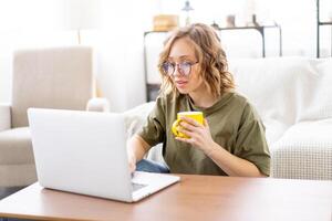 Frau Brille mit Laptop Computer während Sitzung in der Nähe von Sofa mit groß Fenster auf Hintergrund beim Zuhause Innere foto