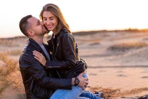 schön jung kaukasisch Paar gekleidet schwarz Leder Jacke und Blau Jeans. ein Mann sitzt auf das Sand Frau sitzt auf oben und Umarmungen ihn. foto