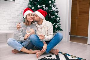 jung glücklich Paar im Weihnachten Hüte in der Nähe von ein Weihnachten Baum küssen, halten Brille von Wein. Neu Jahr Feier foto