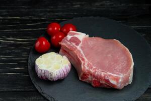 oben Aussicht von einer Stücke roh Schweinefleisch hacken Steaks mit Kirsche Tomaten und Knoblauch auf ein schwarz Stein Schneiden Tafel. foto