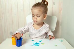 schön wenig Mädchen zeichnet mit Finger Farben auf ein Weiß Blatt von Papier. kreativ Kind Entwicklung im Kindergarten oder kostenlos Zeit beim Zuhause foto