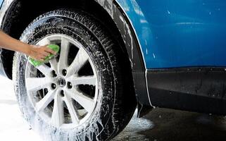 ein Person ist Waschen ein Auto Reifen mit ein Grün Bürste. Reifen ist bedeckt im Wasser und Seife. Blau Auto waschen mit Weiß Seife Schaum. Auto Pflege Service. Auto Reinigung Bedienung Konzept. Fahrzeug Reinigung Service. foto