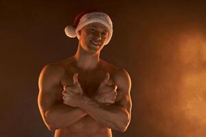 Porträt von muskulös Mann tragen Weihnachten Santa Hut, gefaltet Hände auf rauchig Hintergrund foto