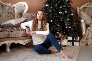 Weihnachten. Frau gekleidet Weiß Sweatshirt und Jeans Sitzung auf das Fußboden in der Nähe von Weihnachten Baum mit Geschenk Box foto