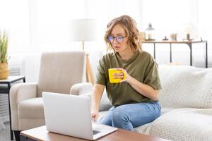 Frau Brille mit Laptop Computer während Sitzung Sofa mit groß Fenster auf Hintergrund beim Zuhause Innere foto