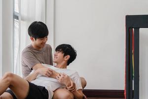 asiatisches schwules Paar, das zu Hause auf dem Boden liegt und umarmt. junge asiatische lgbtq-männer, die sich glücklich küssen, entspannen sich zusammen und verbringen morgens romantische zeit im wohnzimmer mit regenbogenflagge im modernen haus. foto