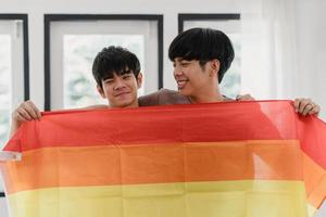 Porträt junges asiatisches schwules Paar, das glücklich ist, Regenbogenflagge zu Hause zu zeigen. asia lgbtq männer entspannen zahniges lächeln und schauen auf die kamera, während sie sich morgens im modernen wohnzimmer im haus umarmen. foto