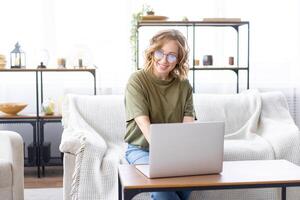 Frau Brille mit Laptop Computer während Sitzung Sofa mit groß Fenster auf Hintergrund beim Zuhause Innere foto