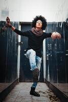 ein jung gut aussehend gemischtrassig afrikanisch amerikanisch steht in der Nähe von das Spiegel Mauer mit ein Fett gedruckt Bein im schwarz Stiefel und Jeans. afro Frisur. Schüler und Jugend Kultur foto