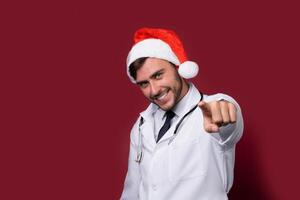 jung gut aussehend Arzt im Weiß Uniform und Santa claus Hut Stehen im Studio auf rot Hintergrund Lächeln und Finger im Kamera foto