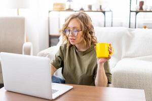 Frau Brille mit Laptop Computer während Sitzung in der Nähe von Sofa mit groß Fenster auf Hintergrund beim Zuhause Innere foto