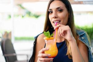 ein schön jung Mädchen von europäisch Aussehen ist Trinken ein exotisch Sommer- Cocktail während Sitzung beim ein Tabelle von ein Straße Sommer- Cafe foto