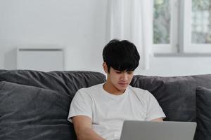 freiberuflicher asiatischer mann, der zu hause arbeitet, männlich kreativ auf laptop auf sofa im wohnzimmer. Geschäftsinhaber Unternehmer, junger Mann, Computer spielen, Social Media am Arbeitsplatz im modernen Hauskonzept überprüfen. foto