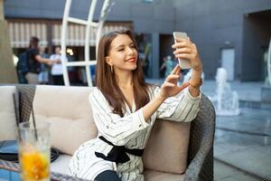 jung lächelnd Frau Herstellung Foto mit Clever Telefon Digital Kamera während Sitzung im draussen Sommer- Restaurant Innere, glücklich weiblich ist Fotografieren Sie selber auf Zelle Telefon während sich ausruhen im Kaffee Geschäft