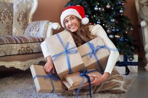 Weihnachten. Frau gekleidet Weiß Sweatshirt Santa Hut und Jeans Sitzung auf das Fußboden in der Nähe von Weihnachten Baum mit Geschenk Box foto