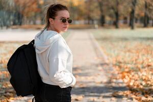 ein jung schön Frau mit ein Pferdeschwanz und Sonnenbrille, mit ein Rucksack auf seine Schultern im das Park. Foto von das zurück