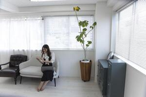 ein japanisch Frau Überprüfung Smartphone durch Fernbedienung Arbeit im das klein Büro foto