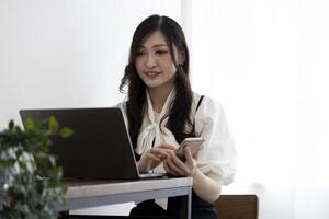 ein japanisch Frau Überprüfung Smartphone durch Fernbedienung Arbeit im das klein Büro foto