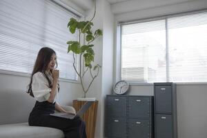 ein japanisch Frau Überprüfung Smartphone durch Fernbedienung Arbeit im das Zuhause Büro foto