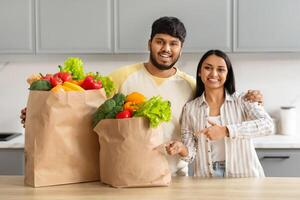 glücklich indisch Paar mit Lebensmittelgeschäft Taschen Stehen im Küche Innere foto