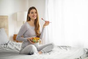 Schwangerschaft Ernährung. schwanger Frau Essen Salat während Sitzung auf Bett beim Zuhause foto