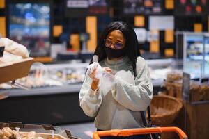 afrikanisch Frau tragen Einweg medizinisch Maske Einkaufen im Supermarkt während Coronavirus Pandämie Ausbruch. Epidemie Zeit foto