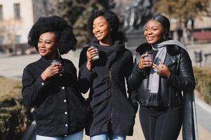 drei stilvoll afrikanisch amerikanisch Mädchen Trinken Kaffee auf das Straße und haben Spaß foto