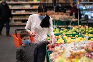 Einkaufen mit Kinder während Virus Ausbruch. Mutter und Kind tragen chirurgisch Gesicht Maske Kauf Obst im Supermarkt. Mama und wenig Junge Kaufen frisch Gemüse im Lebensmittelgeschäft speichern. Familie im Geschäft foto