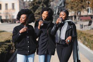 drei schön und stilvoll dunkelhäutig Mädchen mit lange Haar Stehen im ein Stadt und Trinken ein Kaffee und verwenden das Telefone foto