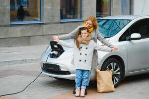 Mutter mit Tochter Laden Elektro Auto beim das elektrisch Gas Bahnhof und sprechen auf Handy, Mobiltelefon Telefon foto