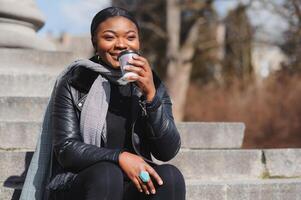 heiter dunkel gehäutet Frau genießen Kaffee halten zu gehen Tasse neu erstellen im Stadt Park, glücklich modisch gekleidet afrikanisch amerikanisch Hipster Mädchen Sitzung draußen während sonnig Tag Trinken Getränk foto