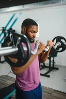 muskulös Mann tun Gewicht Heben Übung beim Fitnessstudio mit Kopieren Raum. Seite Aussicht von passen afrikanisch amerikanisch Kerl Stehen mit schwer Gewicht auf Hantel beim Crossfit Verein. reifen Mann Arbeiten auf Bizeps beim Fitnessstudio. foto