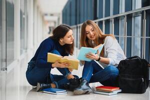 zwei ziemlich weiblich Studenten mit Bücher Sitzung auf das Fußboden im das Universität Flur foto