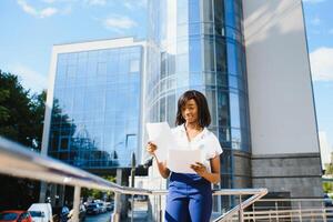 Porträt von jung weiblich afrikanisch amerikanisch Job Sucher behalten ein Mappe mit Lebenslauf im ihr Hände Stehen gegen Büro Gebäude. verschwommen Hintergrund mit Kopieren Raum foto