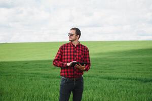 Porträt von Farmer Stehen im jung Weizen Feld halten Tablette im seine Hände und Prüfung Ernte. foto