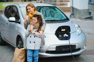 Mutter mit Tochter Laden Elektro Auto beim das elektrisch Gas Bahnhof und sprechen auf Handy, Mobiltelefon Telefon foto